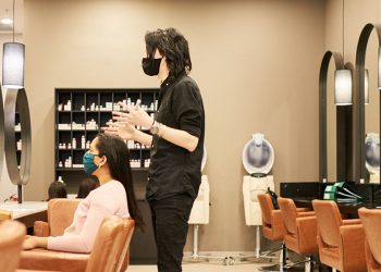 Young male hairstylist wearing a protective face mask talking with a female customer sitting in a chair in his salon
