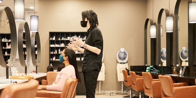 Young male hairstylist wearing a protective face mask talking with a female customer sitting in a chair in his salon