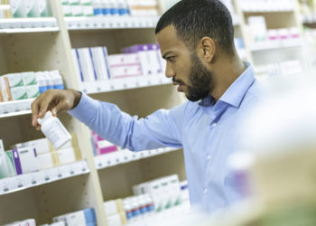 Young African American man standing in front of a shelf at a pharmacy holding bottle of pills and reading instructions.