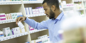 Young African American man standing in front of a shelf at a pharmacy holding bottle of pills and reading instructions.