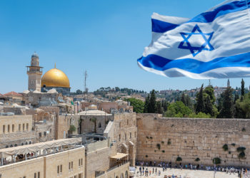 An Israeli flag blows in the wind from an elevated view of the Western Wall. Jewish orthodox believers read the Torah and pray facing the Western Wall, also known as Wailing Wall or Kotel in Old City in Jerusalem, Israel. It is small segment of the structure which originally composed the western retaining wall of the Second Jewish Temple atop the hill known as the Temple Mount to Jews and Christians.
