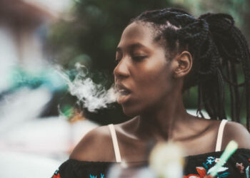 Portrait of a dazzling African girl with braids sitting in a street cafe and exhaling smoke from the hookah; young black female outdoors in a bar plays with the vapor from an electronic cigarette