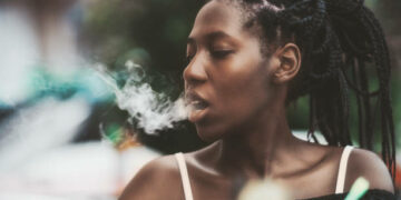 Portrait of a dazzling African girl with braids sitting in a street cafe and exhaling smoke from the hookah; young black female outdoors in a bar plays with the vapor from an electronic cigarette